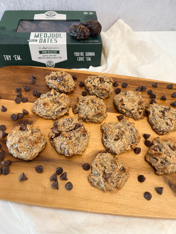 A photo of a wooden platter of Date Sweetened Chocolate Chip Cookies with chocolate chips scattered around the cookies. At the top left is a box of Coachella's Best Organic dates. All on top of a draped cream napkin. 
