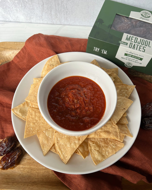 A picture of chips and salsa on a wooden platter with burnt orange napkin draped underneath. A box of Coachella's Best Organic Dates is in the top right corner. 