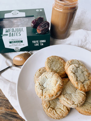 A photo of Peanut Butter Date Cookies piled on a white plate on top of a white napkin. In the background there is a box of dates, a jar of peanut butter, and a spoonful of peanut butter. 