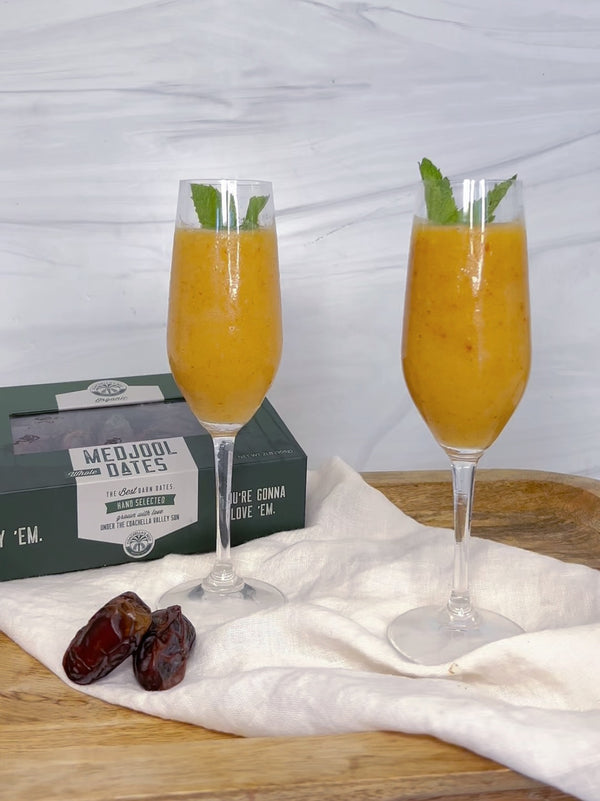 A picture of 2 champagne glasses filled with Peach & Medjool Date Bellini on top of a cream linen napkin draped over a wooden platter. Next to the glasses are 2 medjool dates and a box of medjool dates.