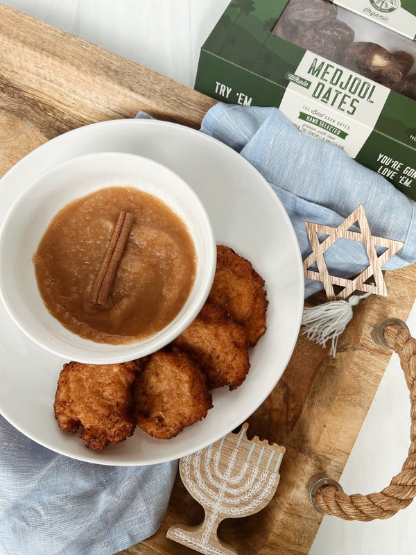 A bowl of Medjool Date Applesauce and a plate of 4 latkes on top of a wooden platter with a blue napkin draped over it. On top of the napkins there are small wooden Hanukkah objects.