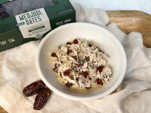 A picture of a bowl of chicken salad with dates on top of a cream napkin draped over a wooden platter. In the top left corner is a green box of Coachella's Best Organic Dates and below the bowl are 2 medjool dates.