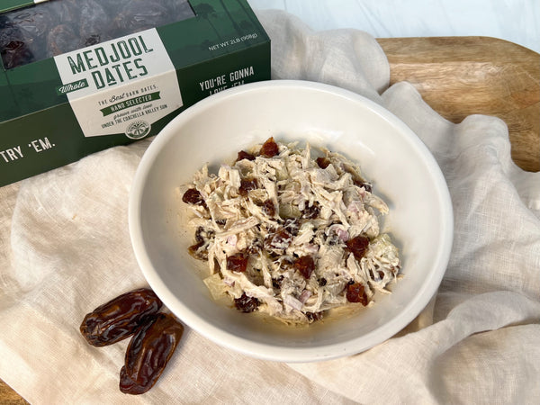 A picture of a bowl of chicken salad with dates on top of a cream napkin draped over a wooden platter. In the top left corner is a green box of Coachella's Best Organic Dates and below the bowl are 2 medjool dates.