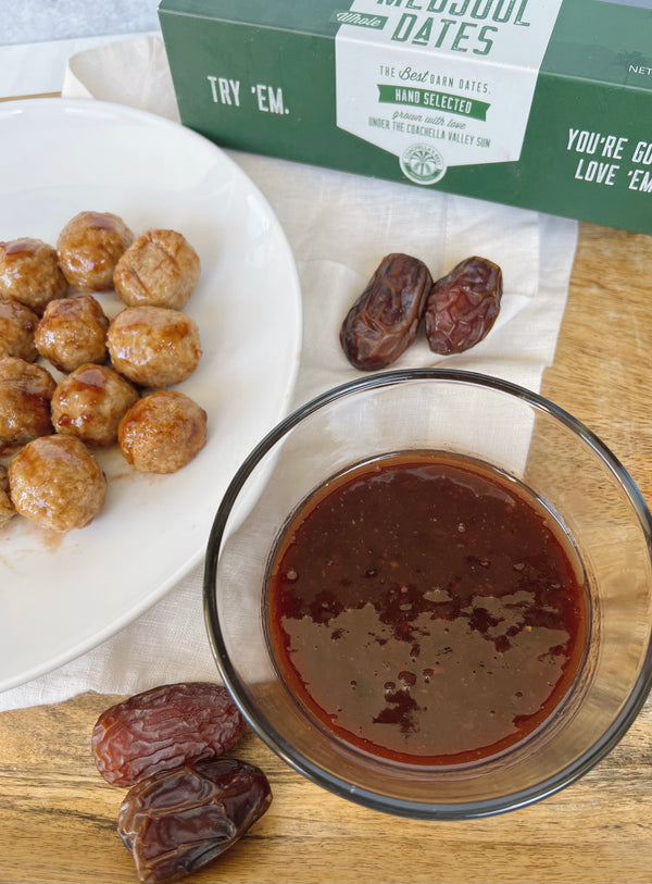 A picture of a plate of meatballs and a bowl of organic balsamic date glaze on a cream linen napkin on top of a light wood platter. At the top there is a cut off box of Medjool Dates and 4 Dates scattered around on the napkin. 
