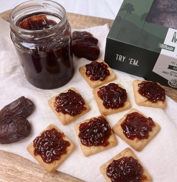 A photo of 8 shortbread cookies with Organic Medjool Date & Berry jam scattered on a cream colored napkin next to a jar of jam. In the corner there is part of the Medjool Date Box showing and also 4 dates scattered around.