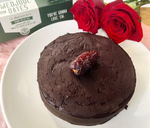 A picture of a full Organic Date Chocolate Cake on a white plate with a box of Medjool Dates and 2 red roses in the background. 