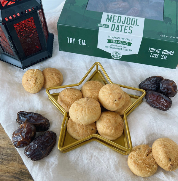 A picture of maamoul cookies in a gold star dish on top of a cream napkin. Surrounding the star dish are dates, maamoul cookies, a Ramadan lantern, and a box of Coachella's Best Medjool dates