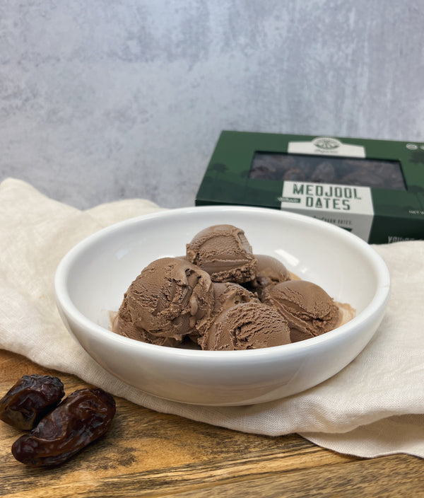 A picture of a white bowl with scoops of Chocolate Date Ice Cream on top of a white clothe draped over a wooden platter. In the background there is a box of Medjool Dates and in front of the bowl are 2 Medjool Dates.