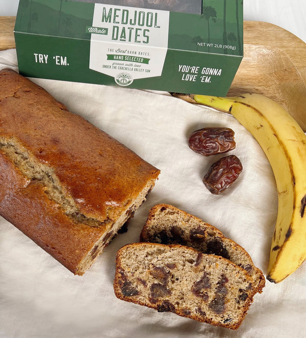 A photo of Banana Bread loaf with 2 slices next to it, a banana, 2 dates, and a box of medjool dates at the top on a wooden board with a cream napkin