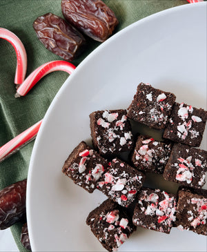 A plate of No Bake Candy Cane Organic Date Brownies on top of a green napkin draped across. On top of the napkin there are a couple of scattered Medjool Dates and 2 full sized candy canes on the side of the plate. 