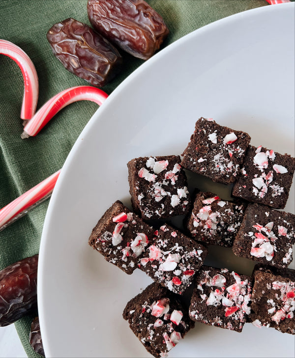 A plate of No Bake Candy Cane Organic Date Brownies on top of a green napkin draped across. On top of the napkin there are a couple of scattered Medjool Dates and 2 full sized candy canes on the side of the plate. 