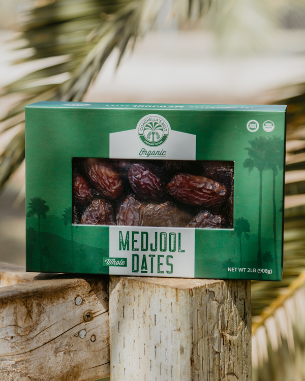 A green 2lb box of Coachella's Best Organic Medjool Dates on top of a wooden peg. Behind are palm tree fronds in the back..
