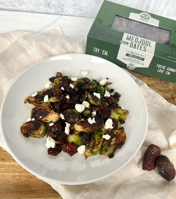 A photo of a plate of Organic Date Brussels Sprouts on top of a cream cloth on top of a wooden platter. At the top right corner is a box of Coachella's Best Organic Dates and on the bottom right is 2 dates piled next to the plate. 