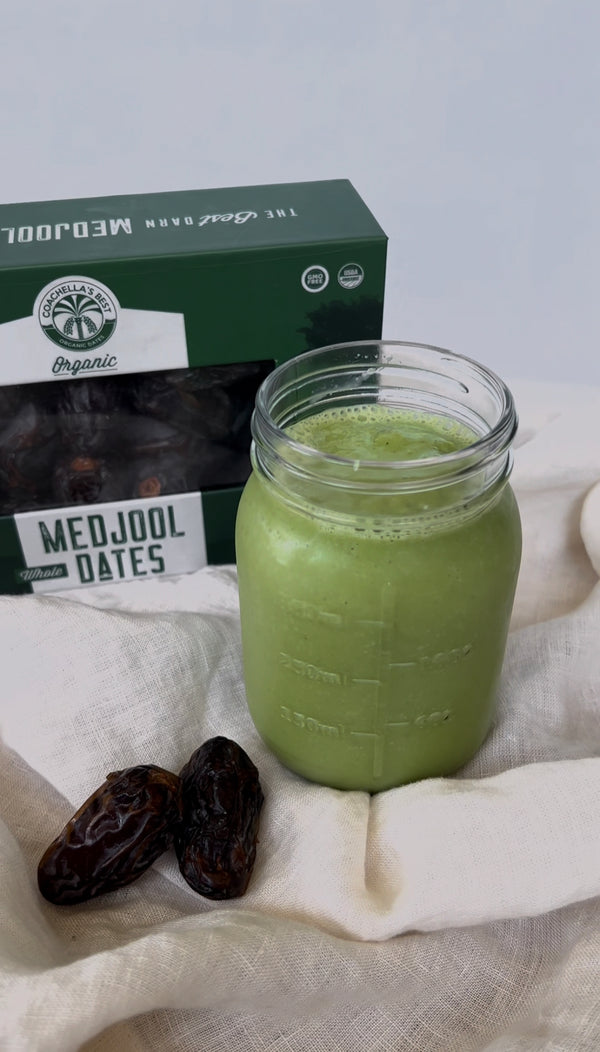 A picture of a Green Date Smoothie on top of a cream linen napkin. Behind the smoothie is a box Coachella's Best Organic Dates and next to the smoothie is 2 dates.